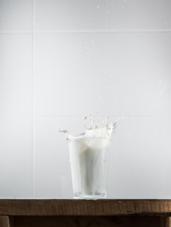 A glass of milk stands on a wooden table in the kitchen. Glass of milk with splashes. The drops fly up. Minimalistic breakfast. Vertical. White kitchen tiles in the backgroundの写真素材