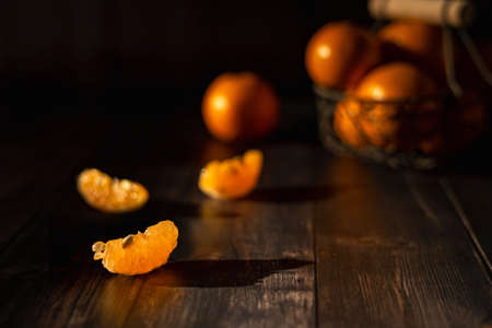 three peeled tangerine slices in the foreground and several tangerines in a basket in the background on a dark tableの写真素材