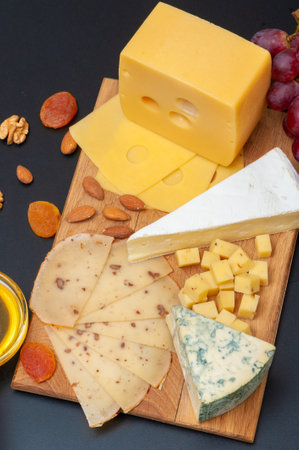 Different types of cheese on a wooden board decorated with honey, grapes, dried apricots and nuts