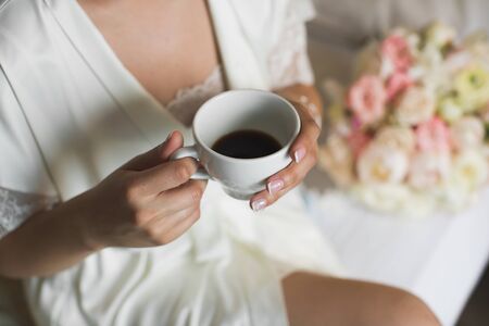 A bride's morning, coffee for breakfast, transparent white peignoir and gentle hands. A picture of an elegant female hand with fine manicure pulling to a perfect cup of coffeeの写真素材
