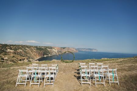 Wedding arch in the form of a ring and white chairs on a cliff against the background of the ocean.の写真素材