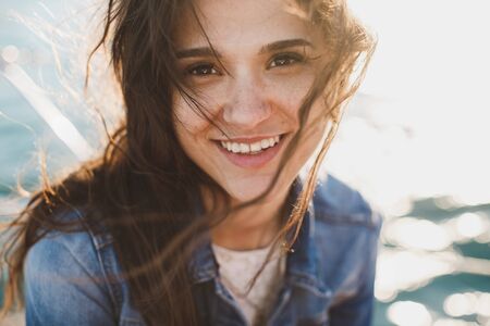 Beautiful young woman at the ocean standing against a turquoise blue sea with her hair blowing in...