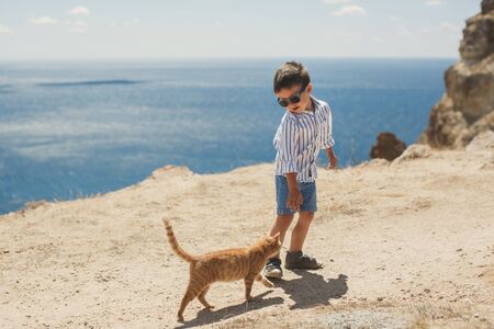 Happy boy playing with red cat against the backdrop of mountains and the sea.の写真素材