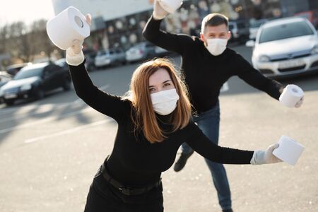 A woman and a man in a coronavirus face mask hold large rolls of toilet paper on a city street and indulgeの写真素材