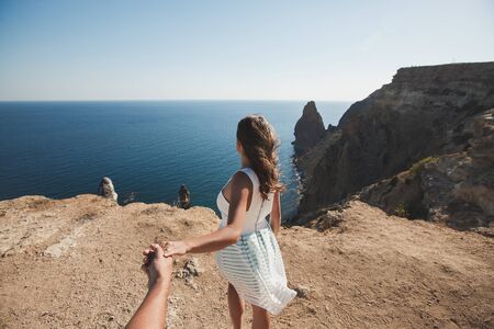 Girl in a white dress stands on a cliff overlooking the forces of the sea and holds the boy by the hand behind the scenesの写真素材