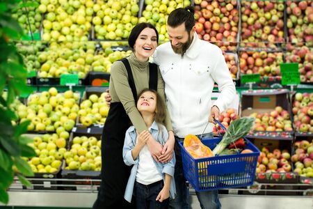 Portrait of a cheerful family standing with a full cart in the supermarket in the vegetable Department on the background of the counter with apples.の写真素材