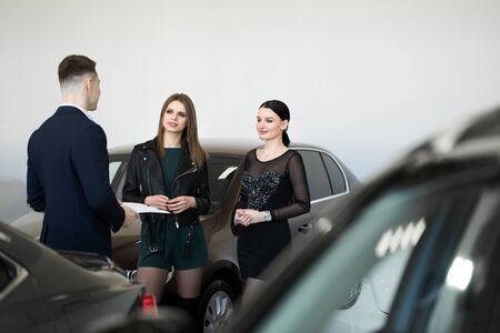 Auto business, car sales - a couple of friends of girls with a car dealer choose a car in a car dealership.の写真素材