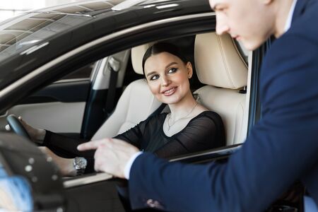 Side view of young beautiful woman sitting inside car and holding hand on steering wheel. She smiling and talking with manager of car dealership. Car agent representing inside of automobileの写真素材
