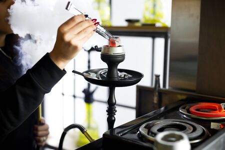 Womens hands hold hookah tongs and adjust the hot coals in a metal bowl. Black hookah stands in a...