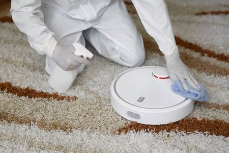 Virologists, people in protective suits carry out the disinfection in the apartment. Wipe the robot vacuum cleaner during the coronavirus epidemic. Covid - 19の写真素材