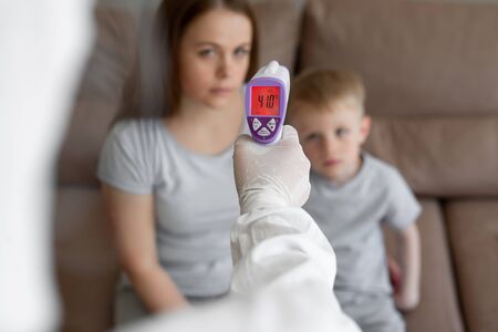 Doctor checks the body temperature of the girl and her son using an infrared thermometer gun on the forehead at home. Coronavirus, covid-19, high fever and coughの写真素材