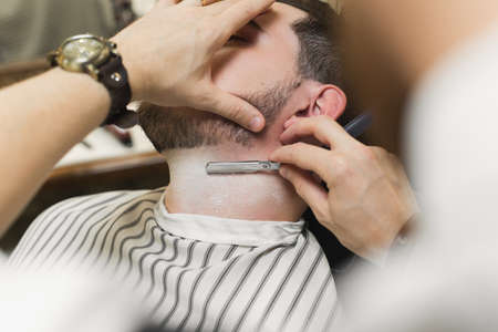 A young man is sitting in a chair in a barber shop while a barber shaves his beard. Preparation of the groom on the wedding dayの写真素材