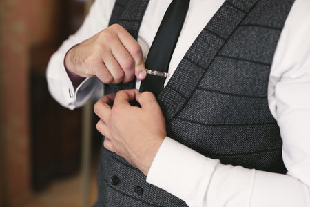 Elegant gorgeous groom getting ready in the morning in the hotel room, holding clip on his tie.の写真素材