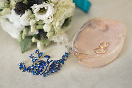 Gold wedding rings on a pink marble shell next to blue jewelry and flowers.