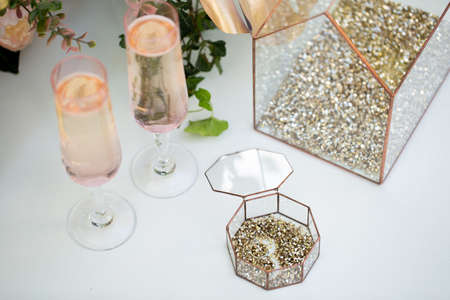Glass box with wedding rings and gold sequins, champagne glasses, flowers on a white table.