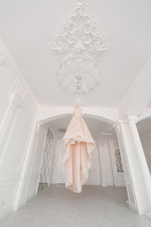 Luxurious peach wedding dress hangs on a chandelier in a white room.の写真素材