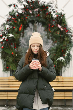 Woman drinks a hot drink on a city street against the background of a cafe window, which is decorated with a Christmas wreathの写真素材