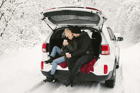 Young couple, a man and a woman, are sitting in the trunk of a car in a winter, snowy forest, hugging, kissing and drinking coffeeの写真素材