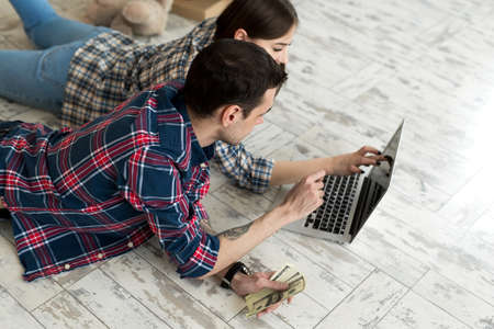 Portrait of a cute young couple lying on the floor at home and managing budget using laptop.の写真素材
