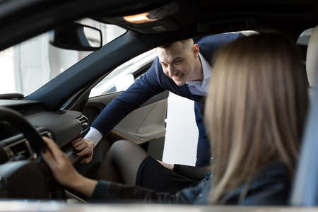 Side view of young beautiful woman sitting inside car and holding hand on steering wheel. She smiling and talking with manager of car dealership. Car agent representing inside of automobileの写真素材