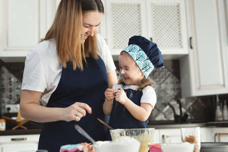 Cute little girl and her beautiful mom in matching aprons and caps play and laugh while kneading...