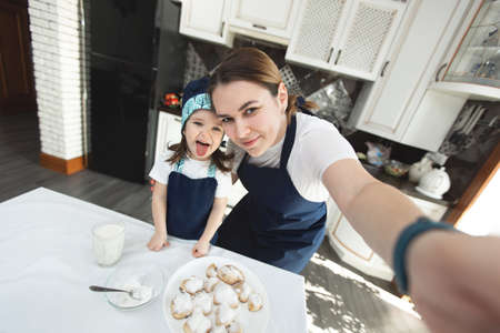 Mom and her daughter bake cookies at home in the modern kitchen and making selfie.
