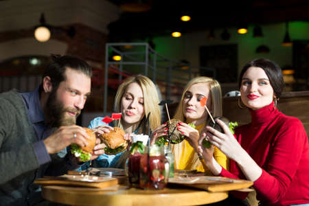 People have dinner together at a table in a cafe. Happy friends eat burgers and drink cocktails in...