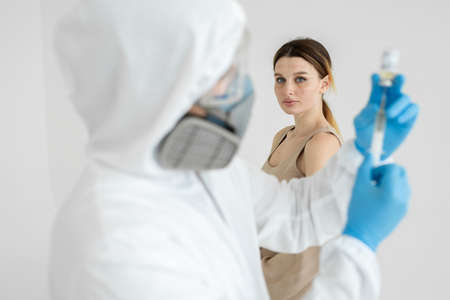 Doctor in a protective suit and mask holds an injection syringe and vaccine COVID-19. Man in a...