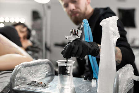 Tattoo master dips a tattoo machine with needle in water. Tattoo artist prepares tools and ink before work in tattoo parlorの写真素材
