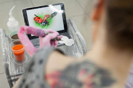 Tattoo master dips a tattoo machine with needles in black ink. The tattoo master applies a colored tattoo on the skin of a young girl, in the background is a tablet with a picture of a ladybug.の写真素材