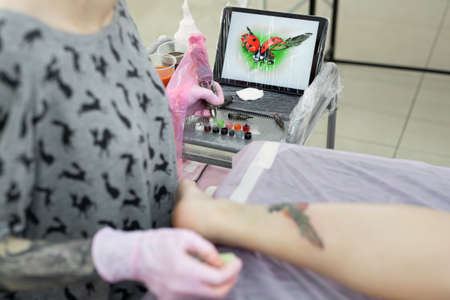Tattoo master dips a tattoo machine with needles in black ink. The tattoo master applies a colored tattoo on the skin of a young girl, in the background is a tablet with a picture of a ladybug.の写真素材