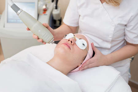 Young woman in safety glasses lying on the couch in a beauty salon, a beautician makes her facial rejuvenation procedure. Close-up of a procedure to rejuvenate the face for a young girl.の写真素材