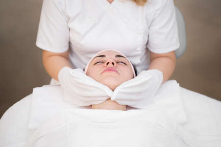 Hands of the beautician wipe the girls face in the cosmetology center with white gloves. Portrait of...