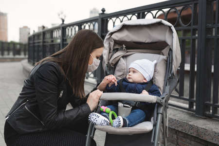 Woman with a mask on her face to protect against coronavirus, covid-19 holds her young child by the...