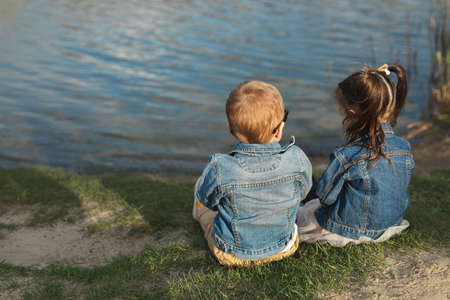Back view of a little boy and girl sitting on the shore of a pond at sunsetの写真素材