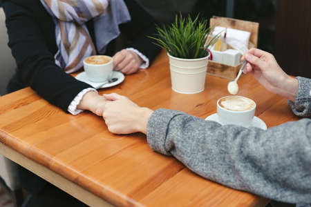 Close-up of a mother and daughters hands on a wooden table in a cafe. Adult mother and daughter hold hands and drink coffeeの写真素材