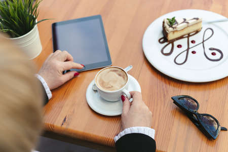 Woman drinks coffee in a cafe on the street. Close-up of a tablet, glasses, coffee and dessert on a wooden tableの写真素材
