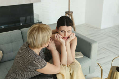 Beautiful young woman is crying on the sofa with her hands on her head. The elderly mother calms her, holds her hand and hugs herの写真素材
