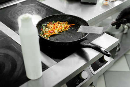 Close-up of the hands of a chef who is frying meat and vegetables in a pan in a restaurantの写真素材