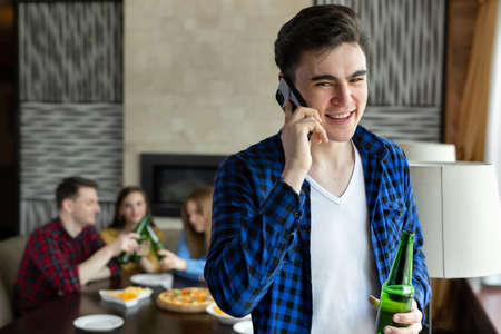 Young man drinks beer from a bottle, talks on the phone and looks out the window of a cafe against the background of his friends who are sitting at the table and eating pizzaの写真素材