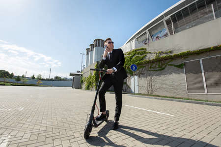 Man in a black business suit stands next to an electric scooter and talks on the phone