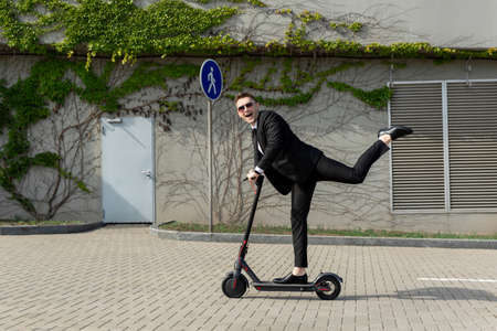 Man in a business suit and sunglasses rides an electric scooter on one and laughs