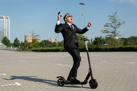 Young handsome businessman in a suit rides an electric scooter and is happy after a conversation on the phoneの写真素材