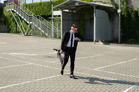 Young businessman in a suit looks at his watch and carries an electric scooter in his handsの写真素材