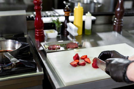 Hand with knife cuts strawberry. Berries on white cooking board. Chef working in the kitchen.