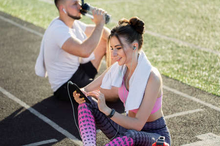 Sporty young boy and woman sit on the treadmill at the stadium in the summer after a run with a phone, headphones and sports bottles.の写真素材