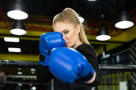 Young beautiful athlete in blue boxing gloves poses in the ring and looks at the camera