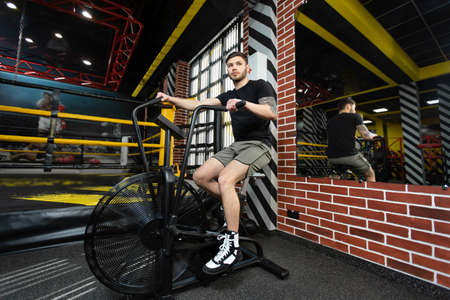 Young athletic male boxer trains on an exercise bike near the ring