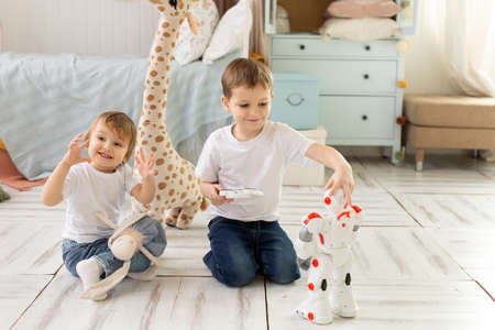 Small children brother and sister sit on the floor in the room, laughing and playing with the robot.