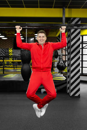 Thin man in a red tracksuit pulls himself up on a crossbar in the gym.